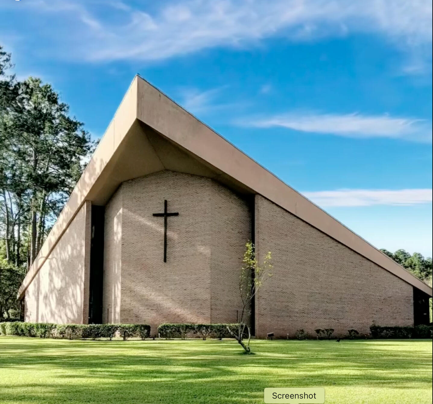 The front of the church building, which is an A-lined, light brick building with a large, simple wooden cross on its front wall. A plush green lawn spreads before it and tall pine trees stand to its left. All these stand before against a bright blue sky.